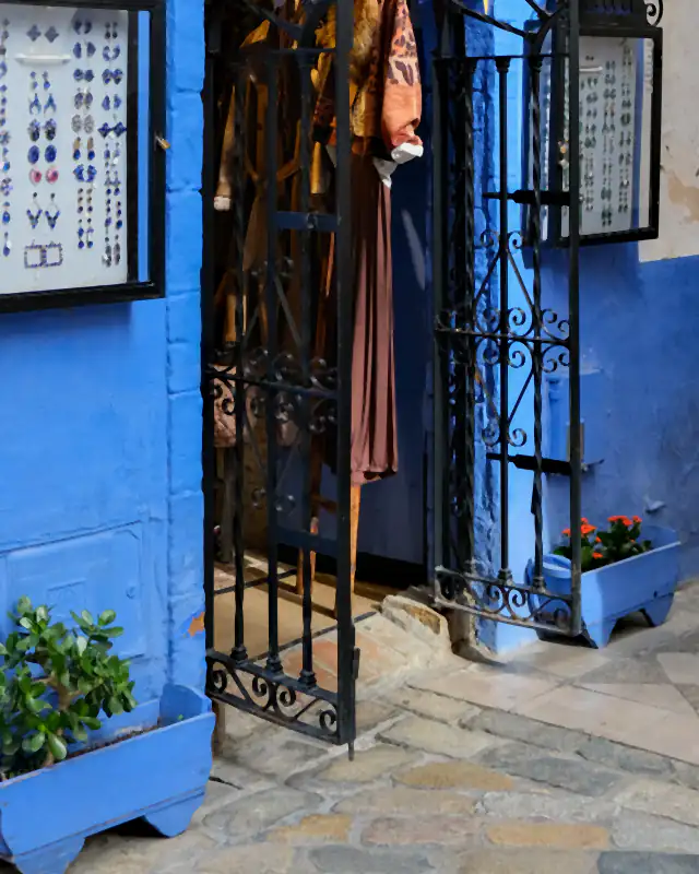 Entrada de una pequeña tienda de barrio en Sevilla: fachada azul, cancela de forja abierta, expositores de bisutería y macetas.